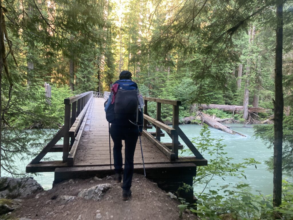 Carolyn walking across a wooden bridge over blue waters of Thunder Creek in North Cascades National Park